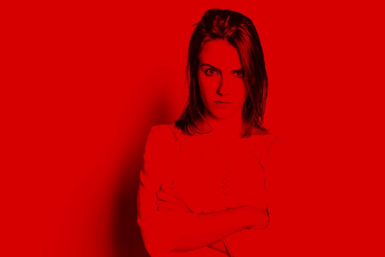 Lit in red, a young woman stands looking angry with her arms crossed.